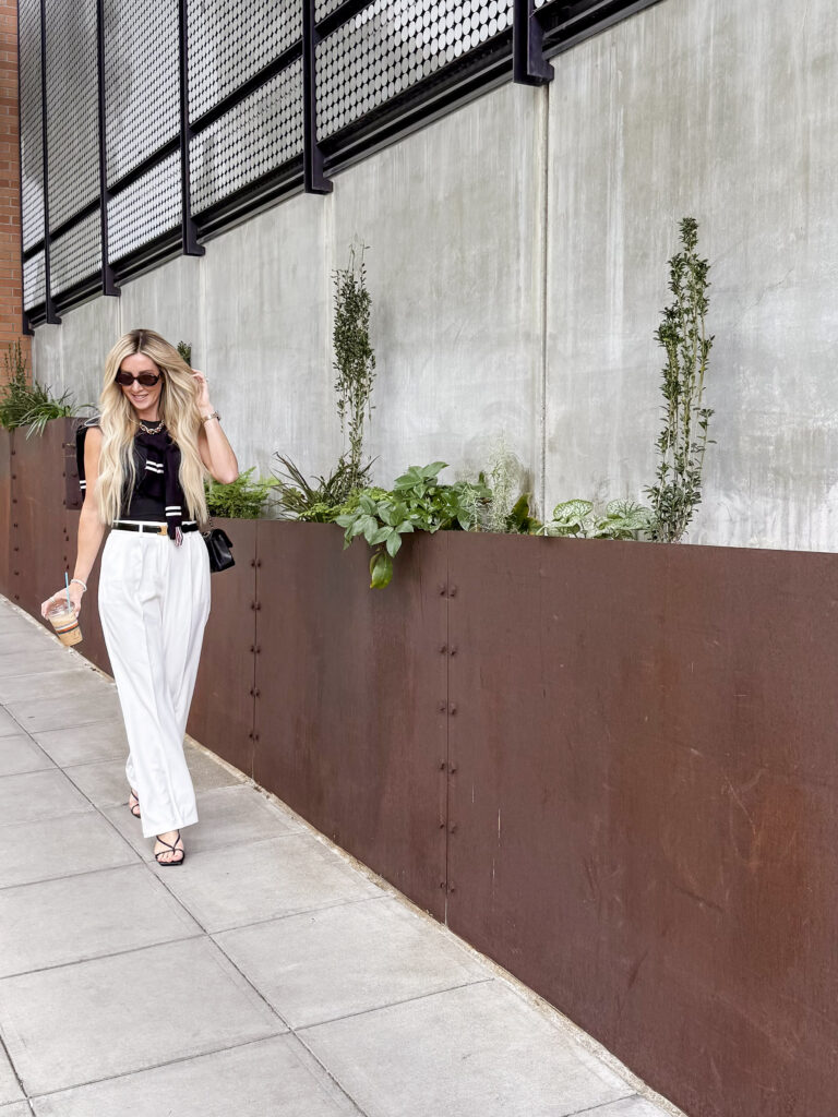 woman walking down the street holding coffee, wearing white pants and a black blouse