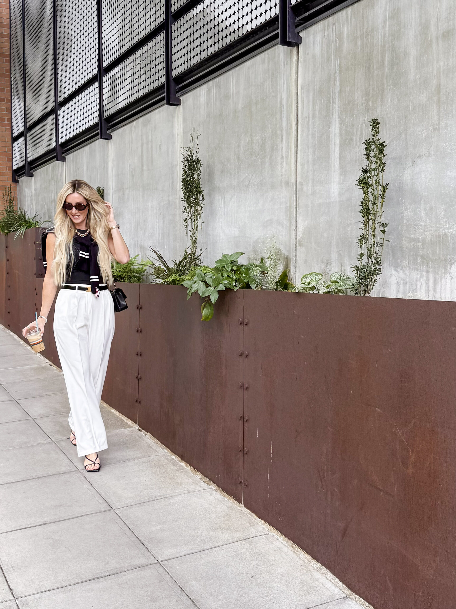 woman walking down the street holding coffee, wearing white pants and a black blouse