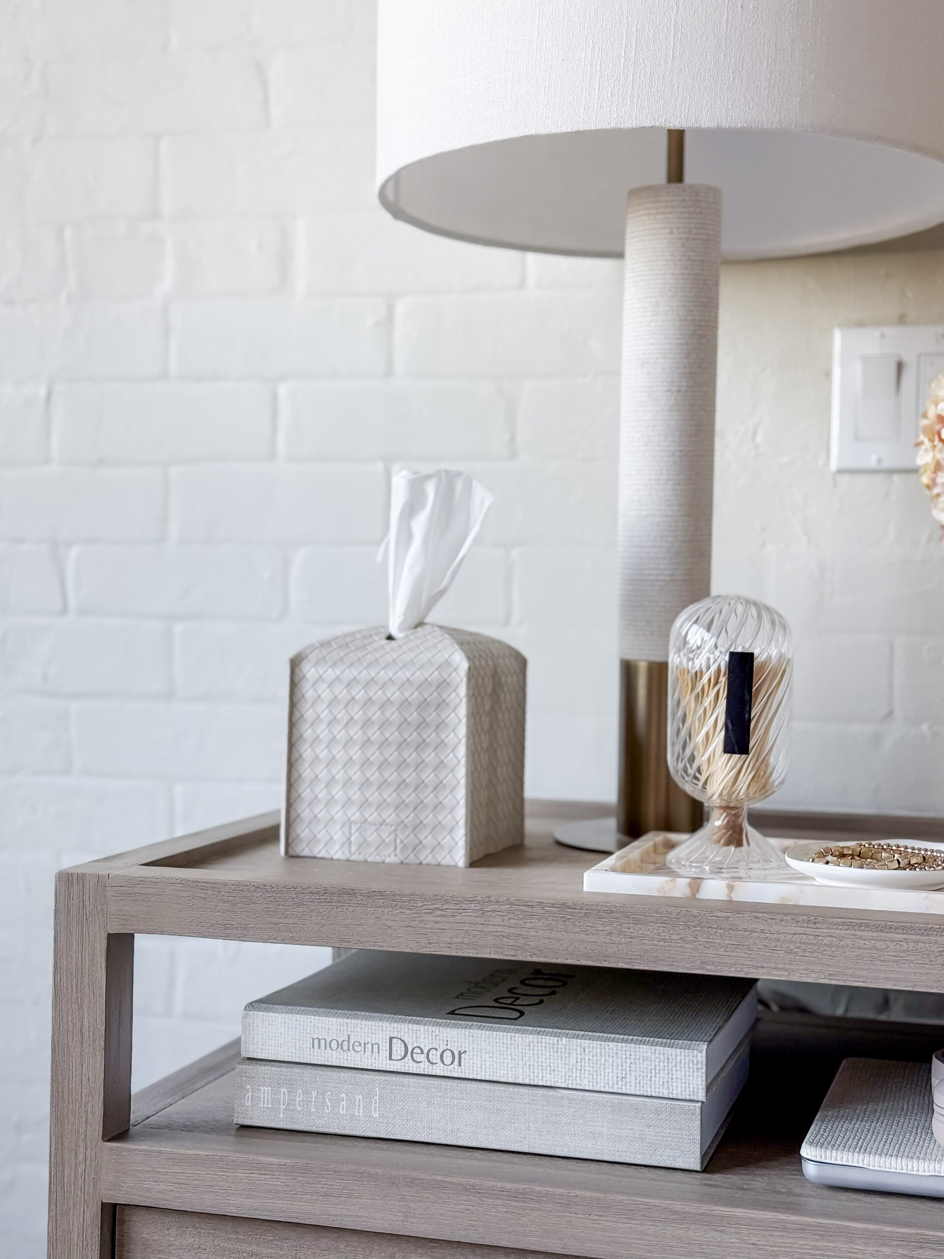 photo of a nightstand with a table lamp, best sellers tissue box cover, match cloche, and decorative books